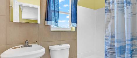Ein Badezimmer mit einem weißen Waschbecken, einer Toilette und einer Badewanne. Ein Fenster mit blauen Vorhängen und ein Spiegel über dem Waschbecken sind vorhanden.