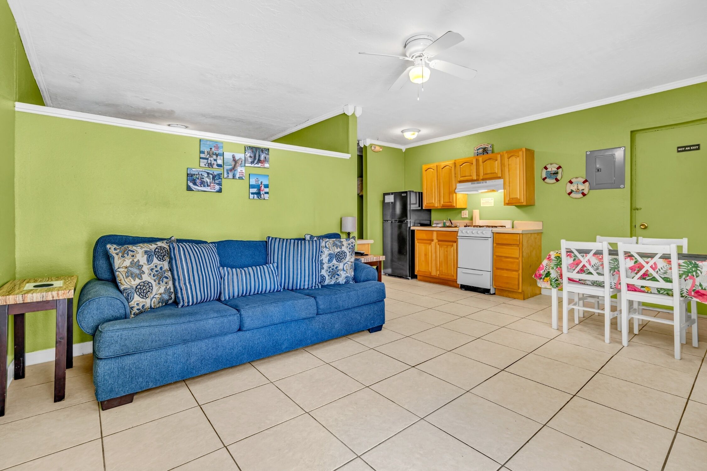 A living room with a blue sofa, a dining table with chairs, a kitchen area with wooden cabinets, and a ceiling fan.