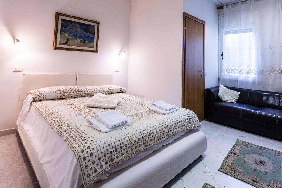A neatly arranged bedroom with a bed, a sofa, and a painting on the wall.
