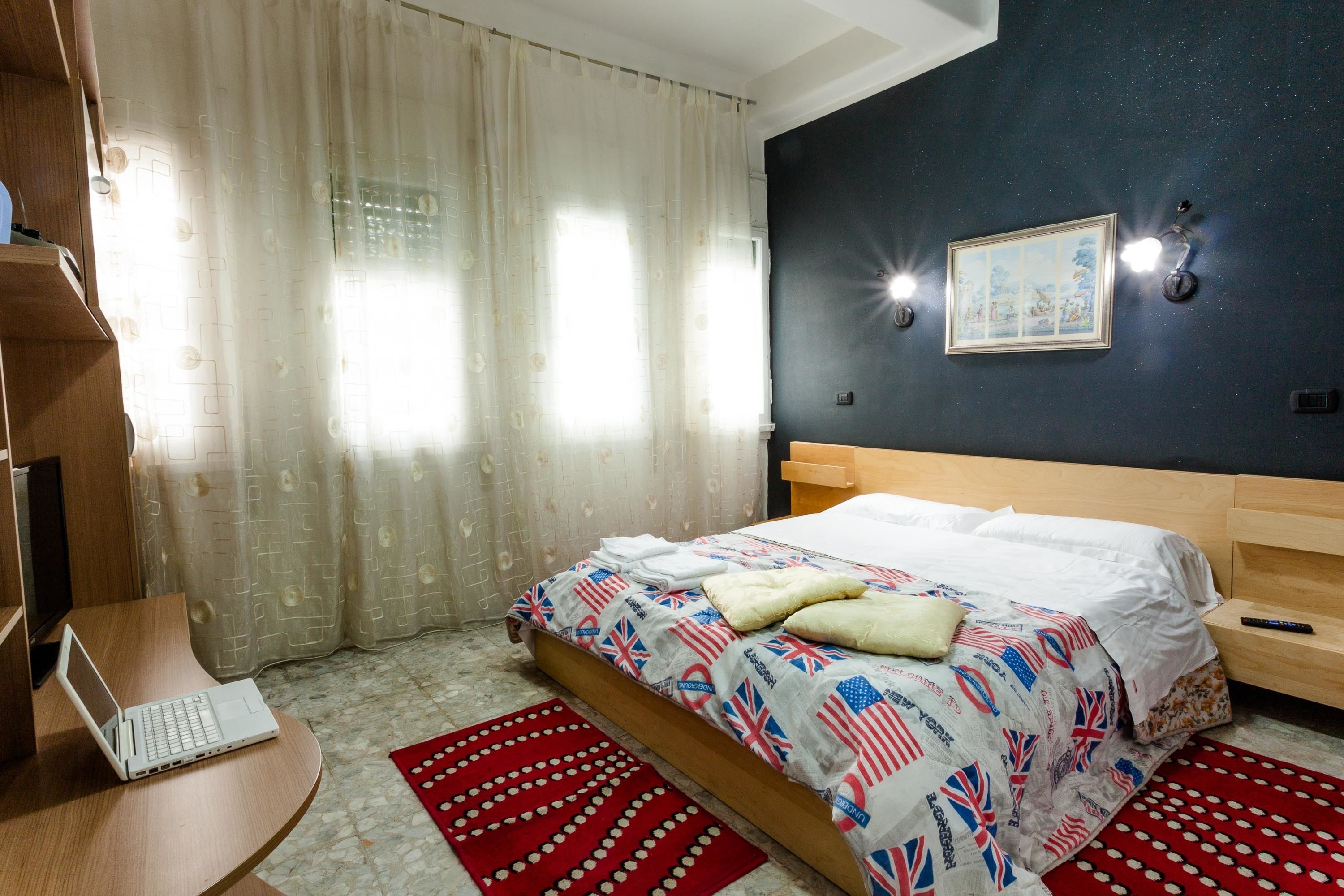 Un dormitorio con una cama, un escritorio con una computadora portátil, un tapete rojo estampado y un cuadro en la pared.