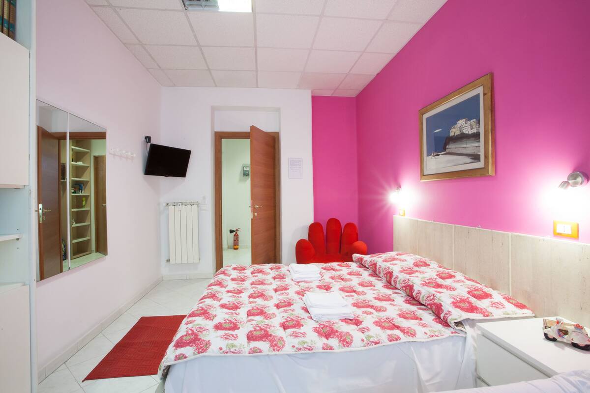 A bedroom with a pink wall, a bed with floral bedding, a red chair, a white bedside table, and a mounted TV.
