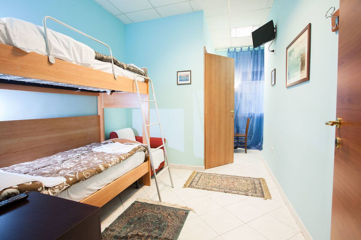 A dormitory room with bunk beds, a desk, a chair, a ladder, and a door leading to another room.