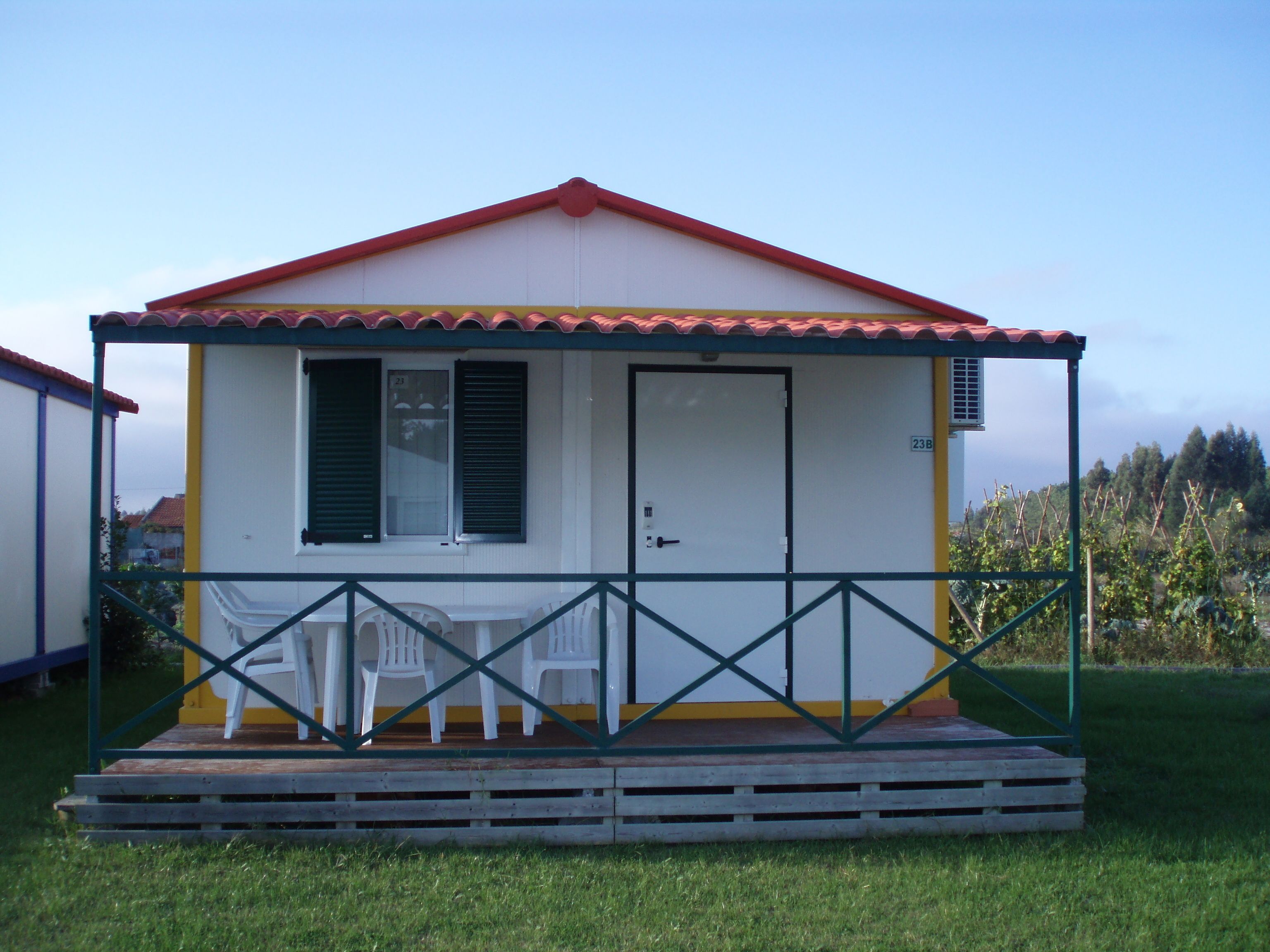 A small, single-story house with a red roof and a porch. There are two windows with shutters, a door, and a small air conditioning unit on the side. The porch has a simple railing and a few plastic chairs.