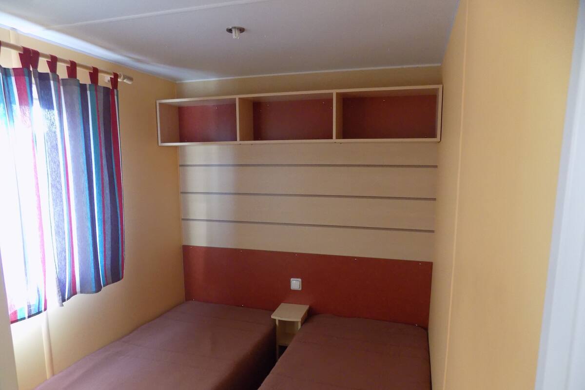 Ein Zimmer mit zwei Betten, einem Fenster mit Vorhängen und einem Schrank über den Betten.