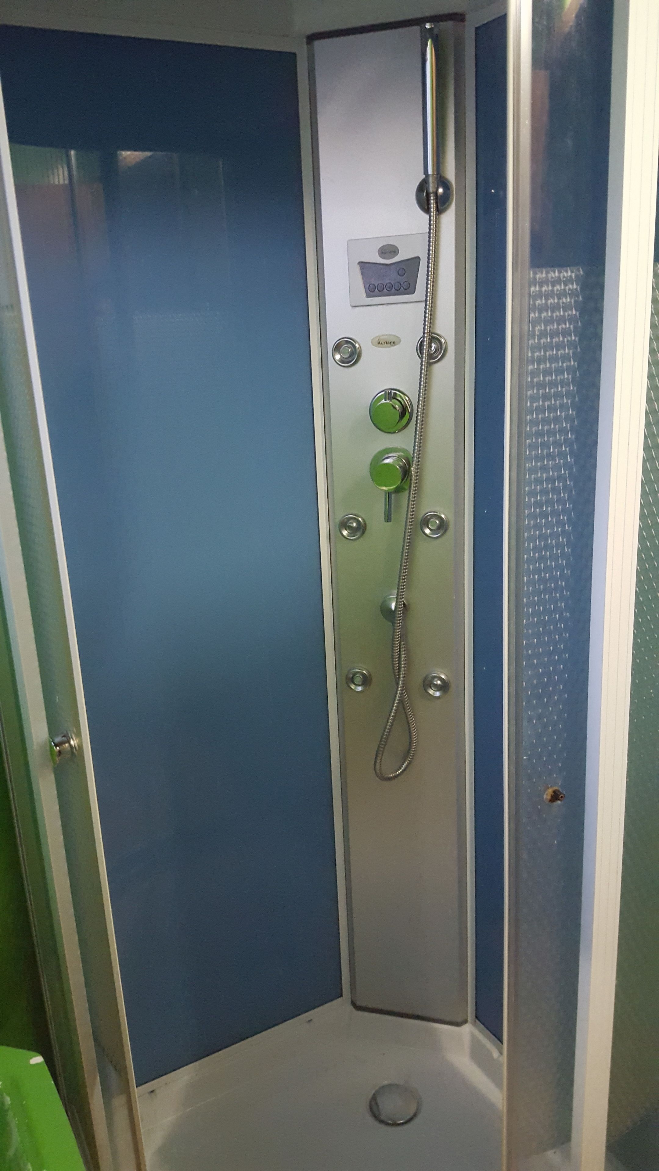 En liten, avskild duschkabin med en kontrollpanel, ett duschmunstycke som kan hållas i handen och ett avlopp i botten.