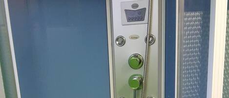 En liten, avskild duschkabin med en kontrollpanel, ett duschmunstycke som kan hållas i handen och ett avlopp i botten.