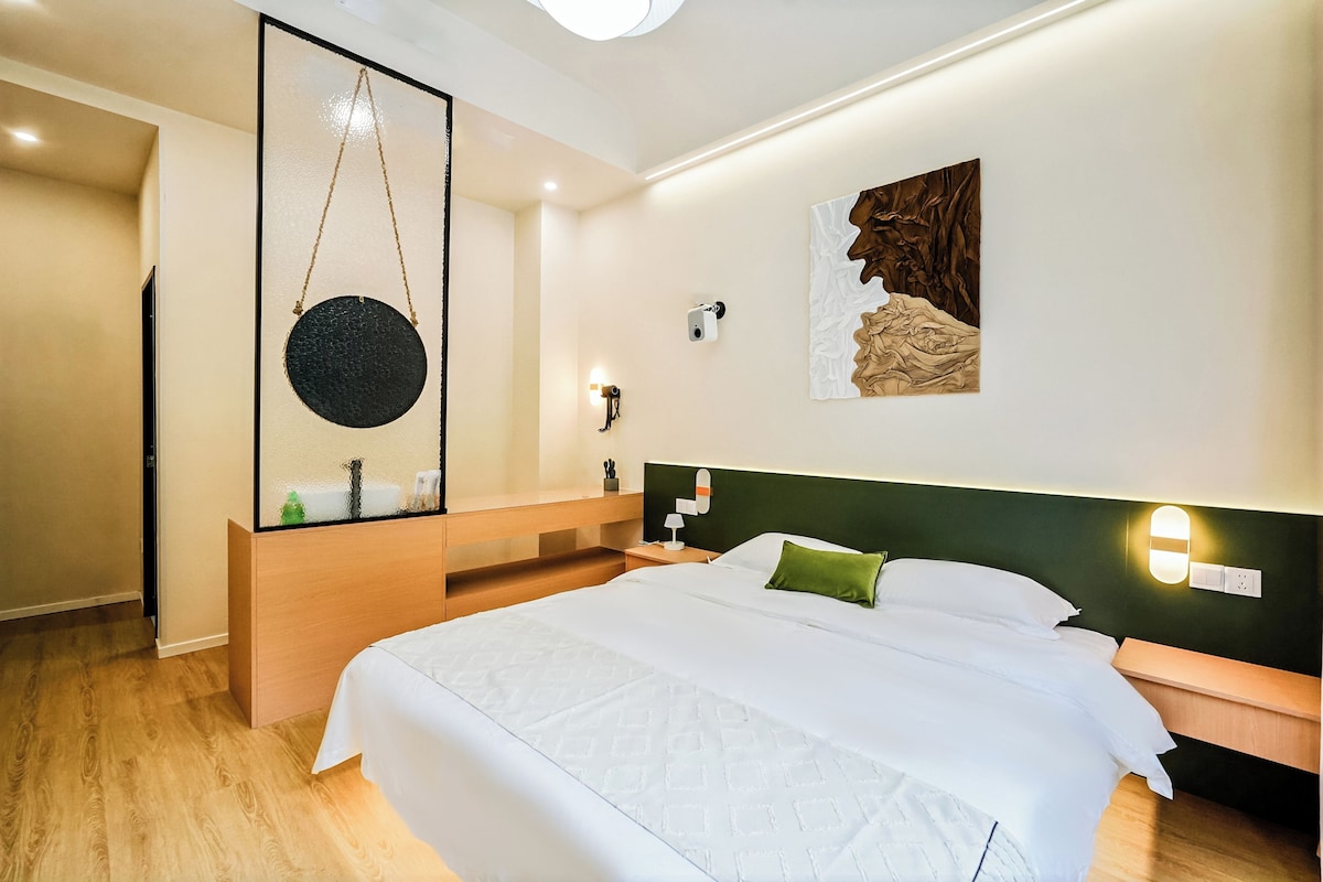 A hotel room with a large bed, a wooden bench, a wall-mounted camera, and a painting.