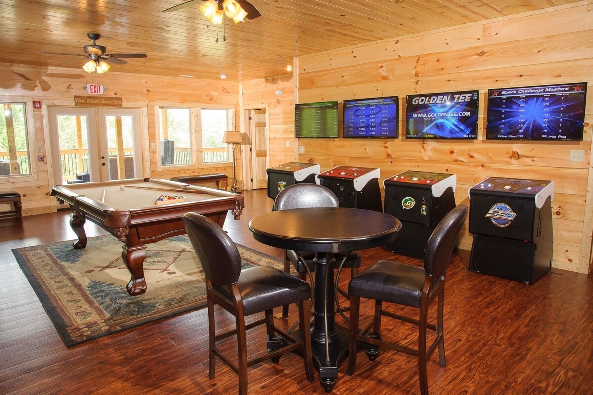 A pool table with four chairs, a dartboard, and three arcade machines in a wooden room with windows.