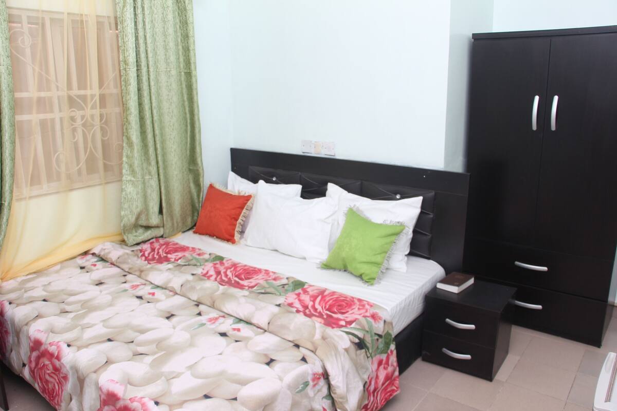 Un dormitorio con cama, almohadas, mesita de noche, armario y ventana con cortinas.