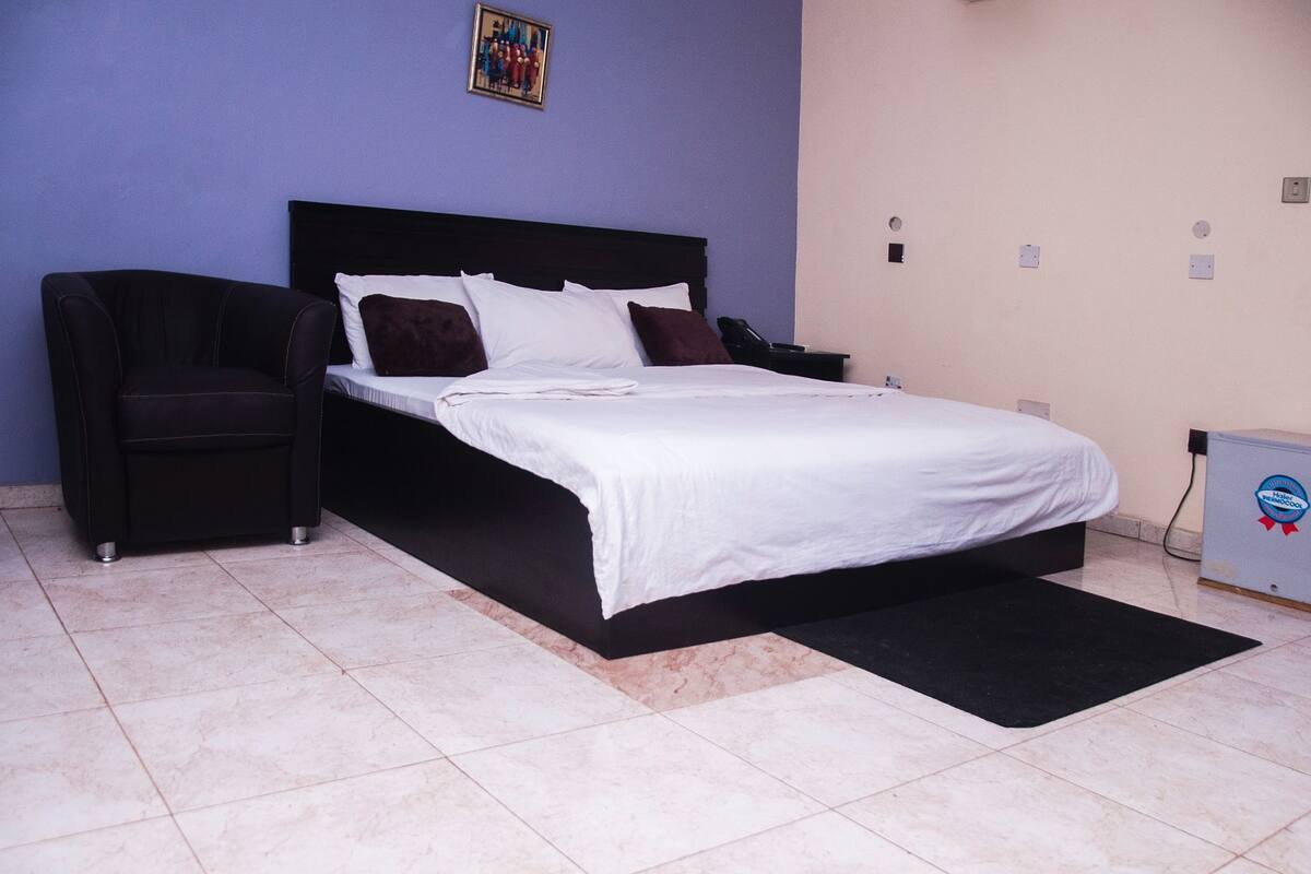 A bedroom with a bed, a chair, a nightstand, and a wall-mounted phone.