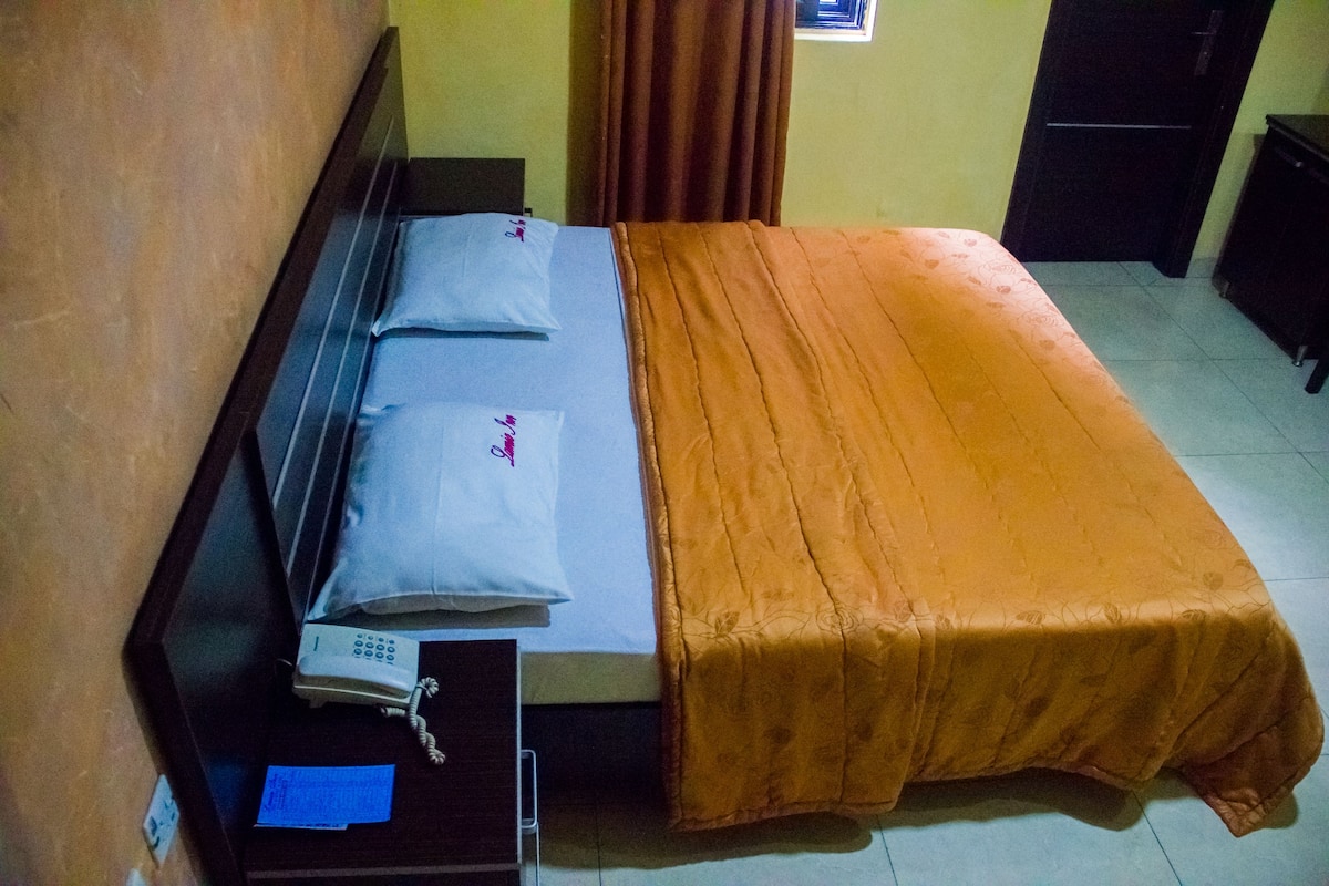 Un lit simple avec un oreiller bleu et une couverture orange, une table de chevet avec un téléphone et une feuille de papier, et une porte en arrière-plan.
