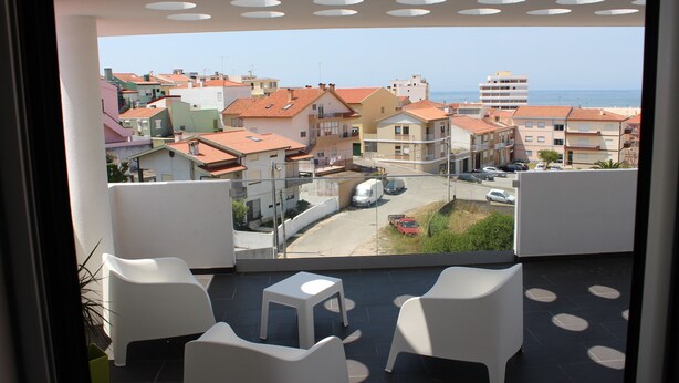 Un balcón con sillas blancas y una pequeña mesa, con vista a una zona residencial de edificios coloridos y una vista despejada del mar.