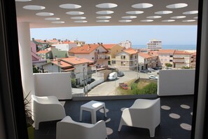 Un balcón con sillas blancas y una pequeña mesa, con vista a una zona residencial de edificios coloridos y una vista despejada del mar.