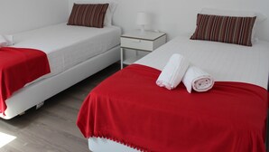 Habitación de hotel con dos camas individuales, cada una con una manta roja y una almohada. Hay una mesita de noche blanca con una lámpara y un cajón pequeño.