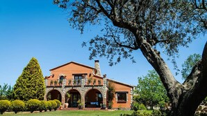 Una casa de dos pisos con fachada de piedra, ventanas arqueadas y un gran árbol en primer plano.