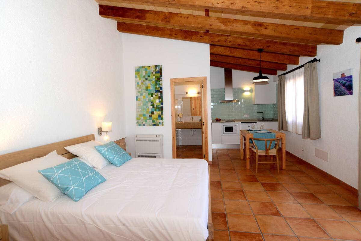 A bedroom with a bed, a wooden ceiling, a small kitchen area with a table and chairs, and a bathroom visible through an open door.