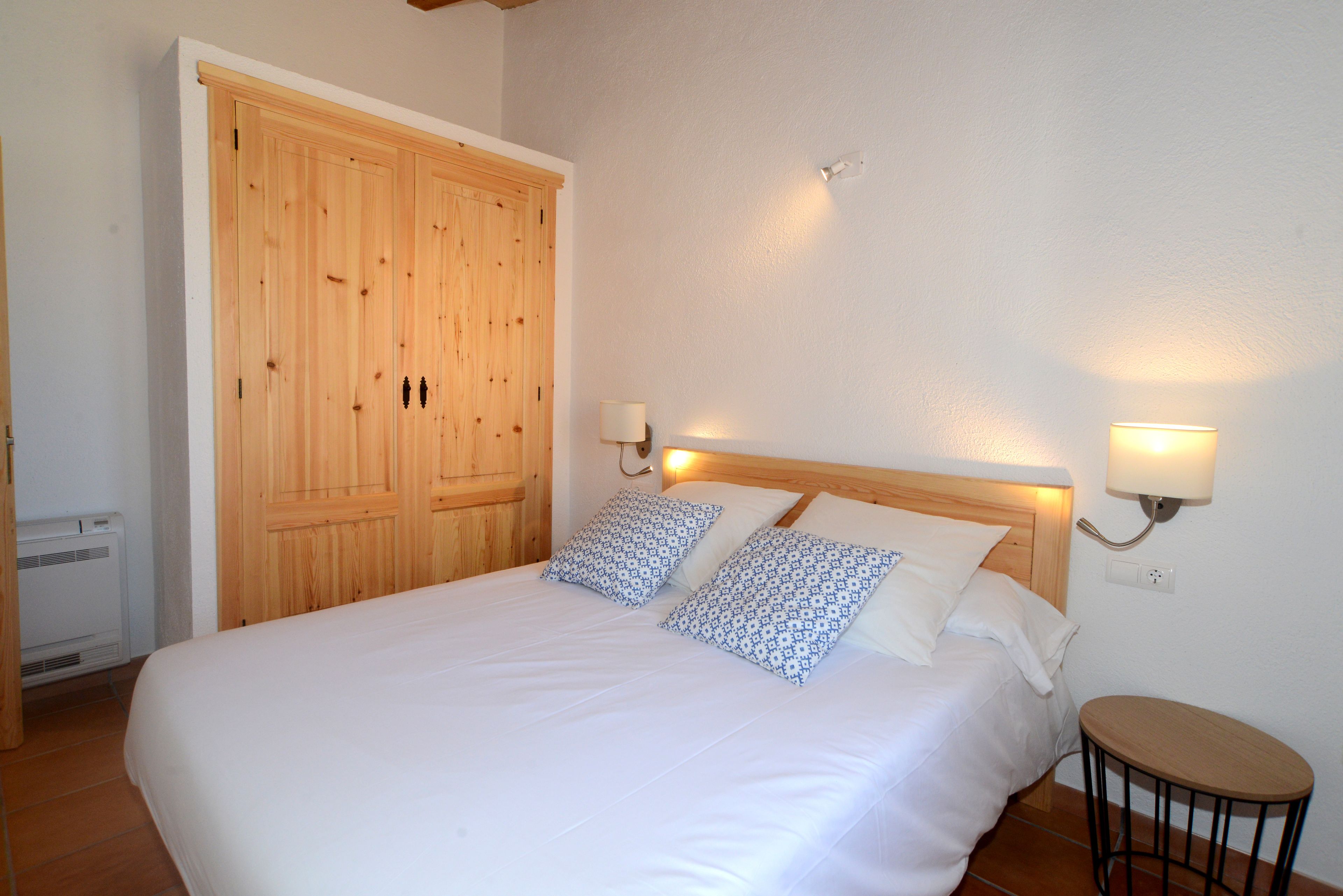Ein Schlafzimmer mit einem Holzschrank, einem Bett mit weißer Bettwäsche, zwei Wandleuchten und einem kleinen runden Tisch.