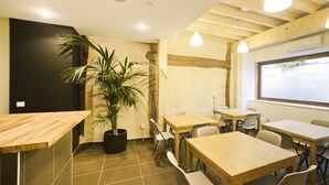 A modern cafe interior with wooden tables and chairs, a large potted plant, and a window with a view. - El Albergue de la Catedral - Hostel (Vitoria-Gasteiz)