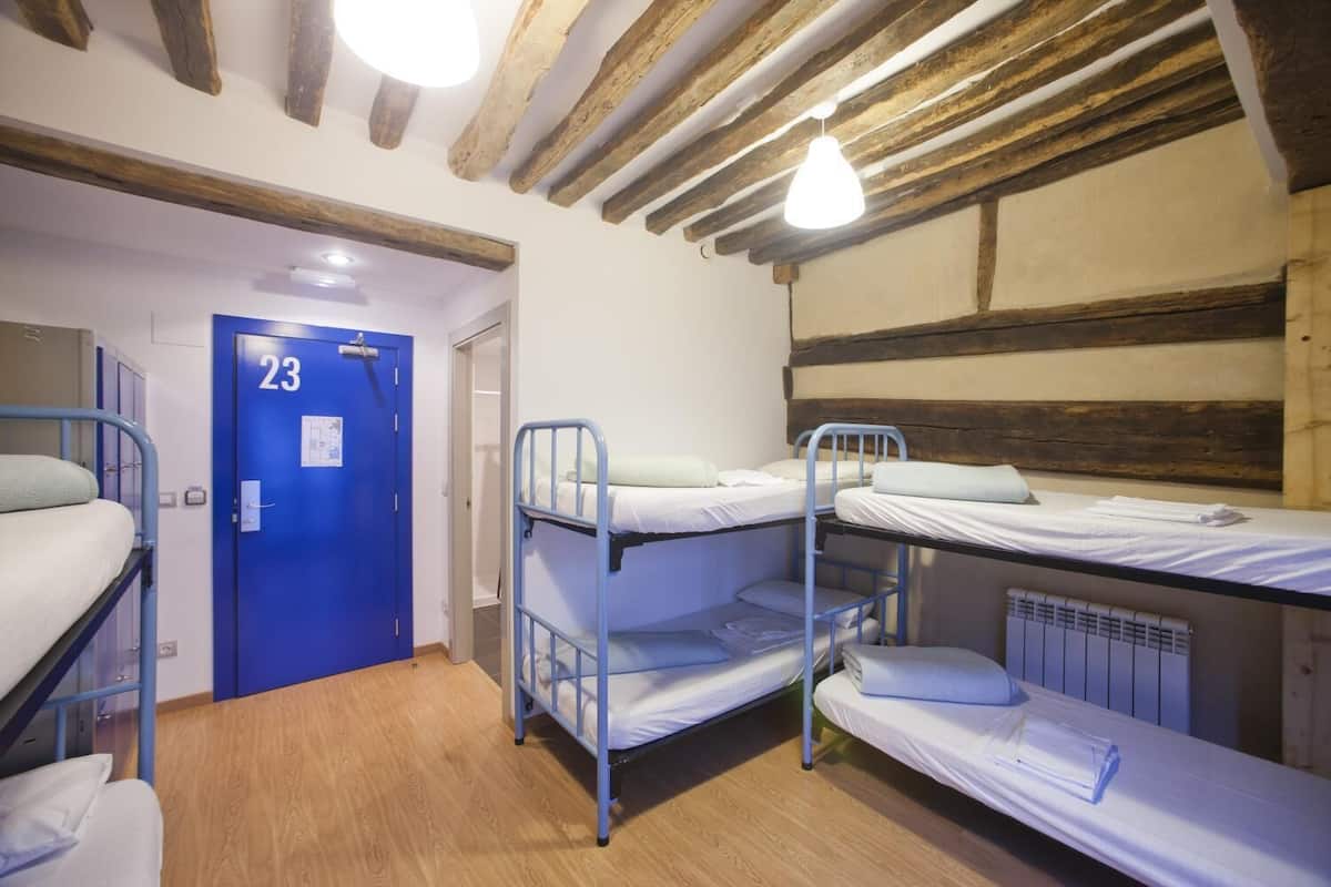 A dormitory room with bunk beds, a blue door with the number 23, wooden beams on the ceiling, and a radiator.