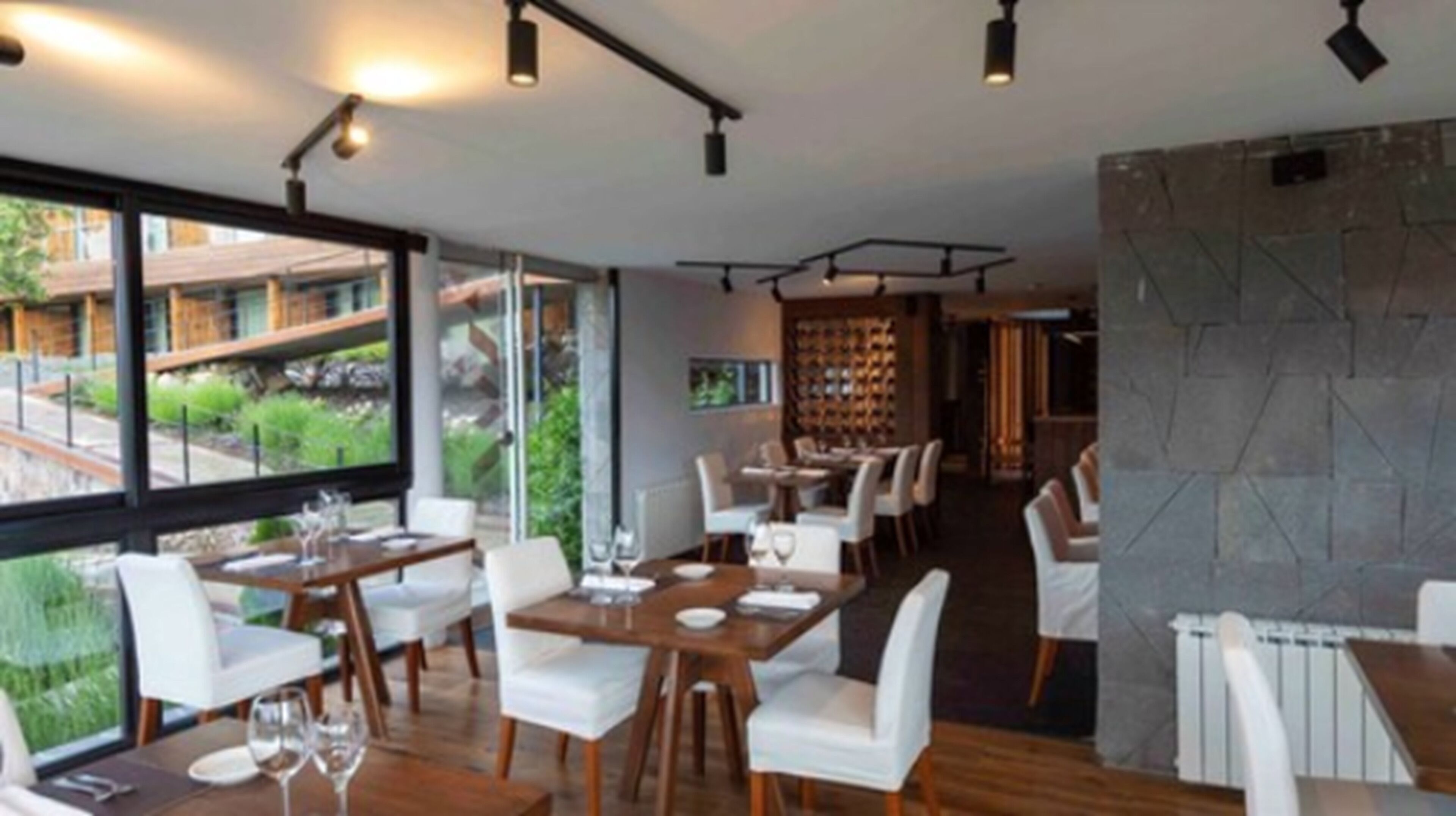A modern dining area with wooden tables and white chairs, large windows offering a view of the outdoors, and a stone wall on one side.