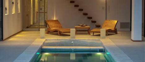 A modern indoor pool area with a glass-enclosed jacuzzi, two wooden lounge chairs, and a staircase leading to an upper level.