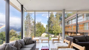 A modern living room with large windows, a sofa, a coffee table, and a view of a lake and mountains.