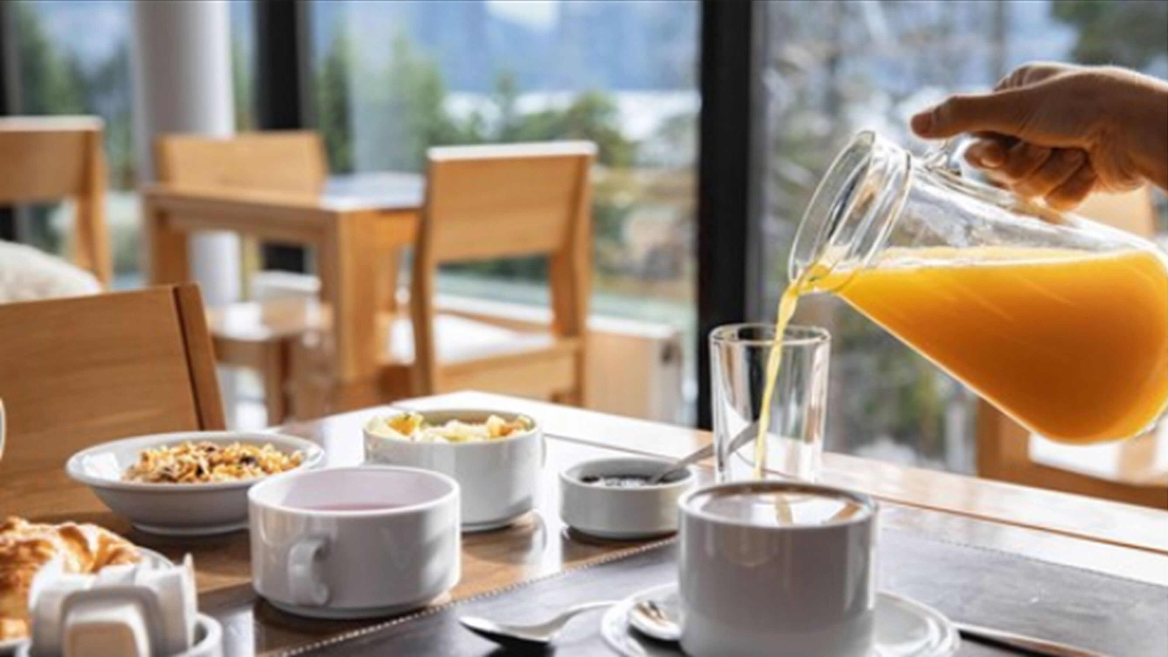 A breakfast setting with a glass of orange juice being poured, a bowl of cereal, and a cup of coffee on a table.