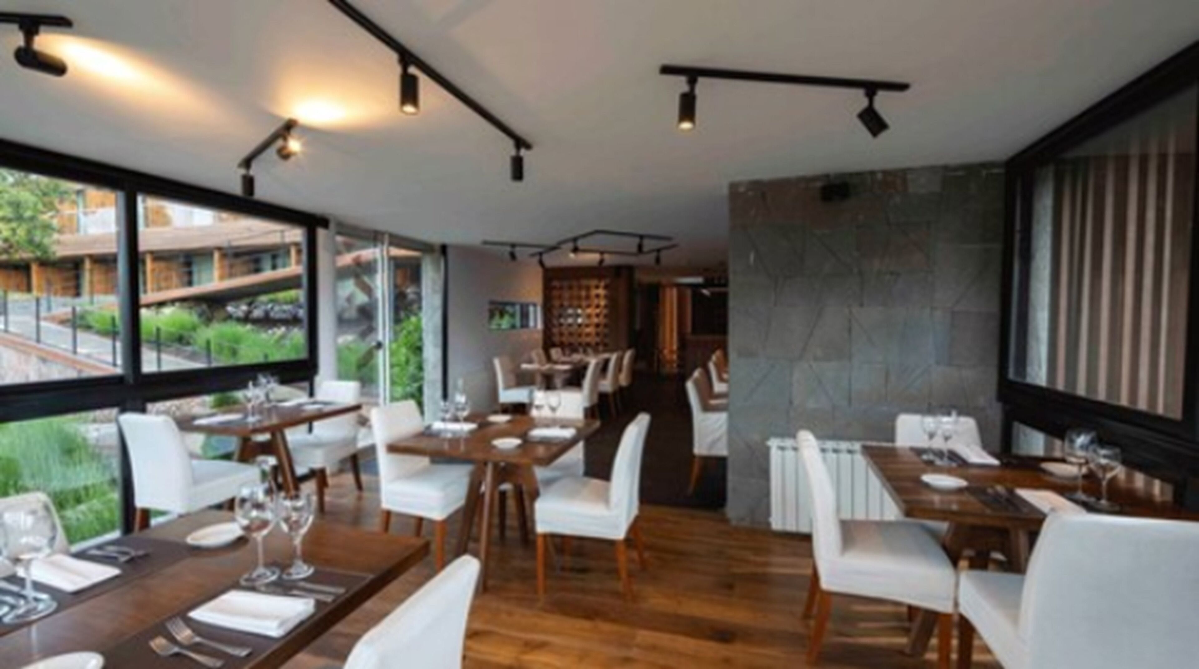 A modern restaurant with wooden tables and white chairs, large windows offering a view of the outdoors, and a stone accent wall.