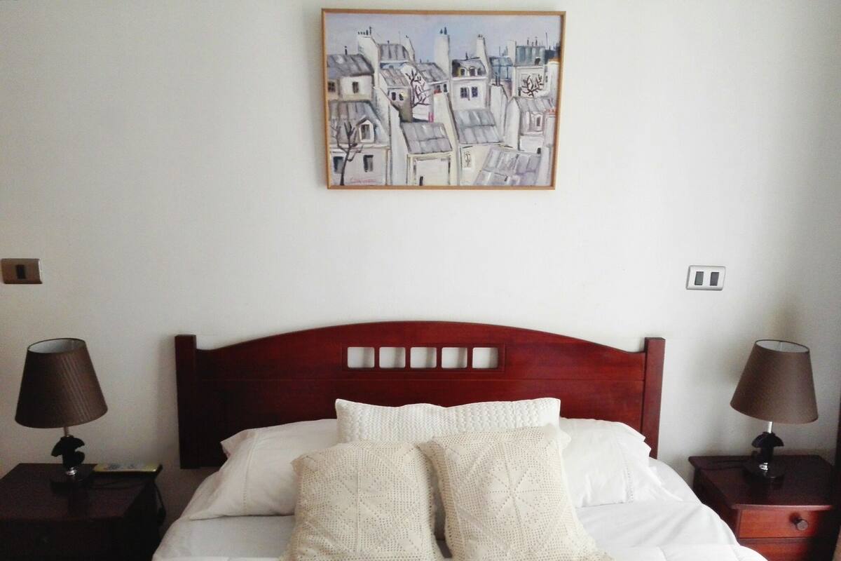Una cama bien hecha con sábanas blancas y dos almohadones decorativos, flanqueada por dos mesitas de noche con lámparas, en una habitación de diseño sencillo y limpio.