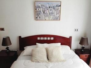A neatly made bed with white linens and two decorative pillows, flanked by two bedside tables with lamps, in a room with a simple, clean design.