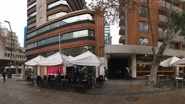 Un área de café al aire libre con mesas y sillas, un edificio de varios pisos y un árbol en primer plano.