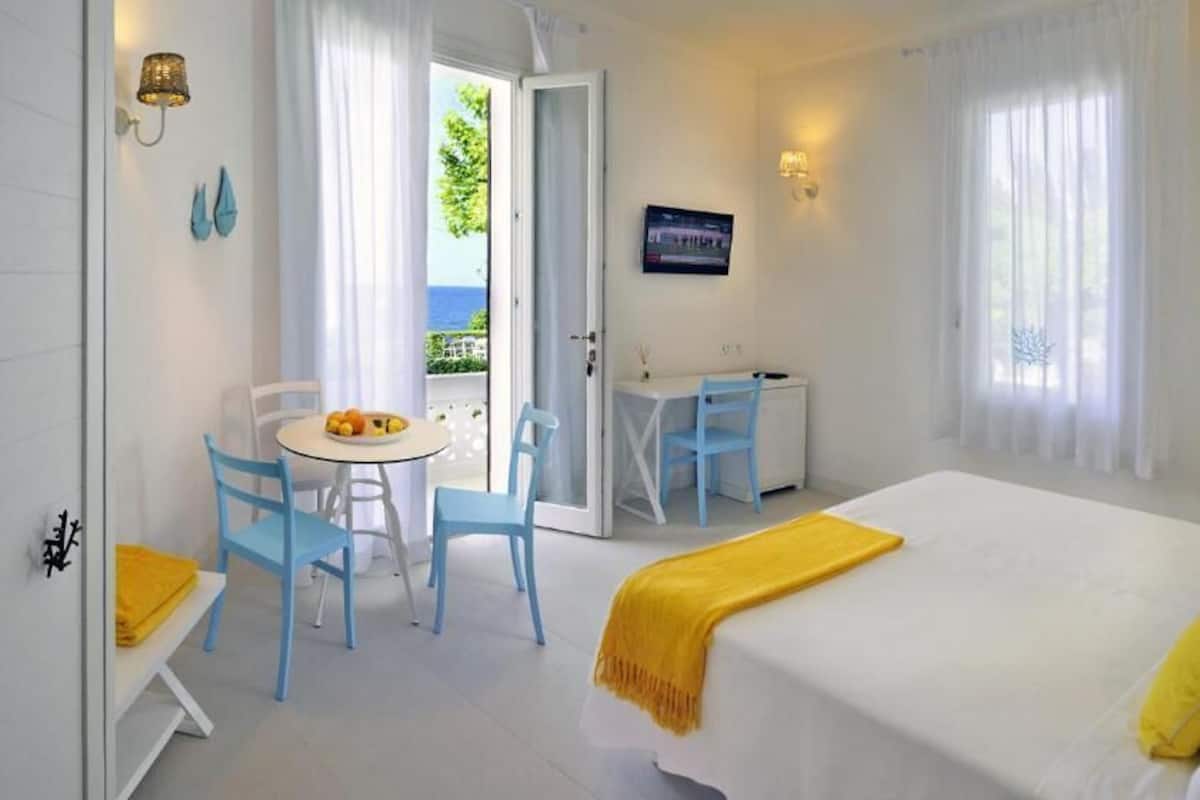 A hotel room with a bed, a desk, and a chair. There is a window with curtains, a TV on the wall, and a view of the outdoors.
