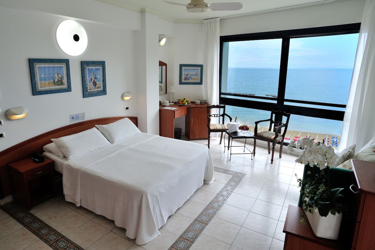 A hotel room with a bed, desk, chair, and a large window with a view of the sea.