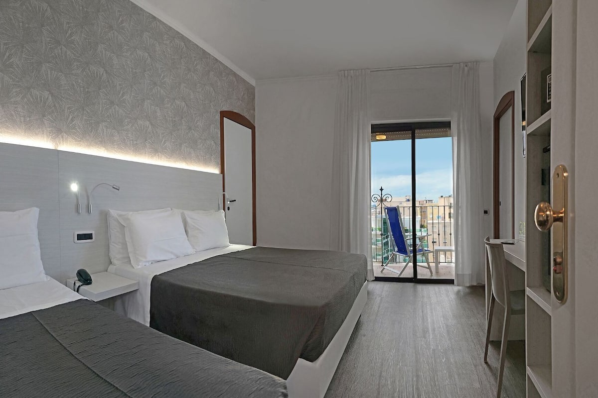 A hotel room with two beds, a bedside table with a phone, a wall-mounted light, and a view of the outside balcony.