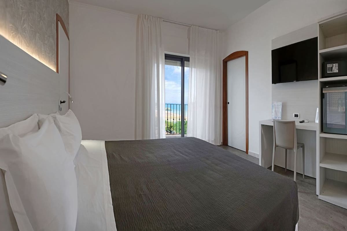 A modern hotel room with a bed, a desk, a TV, and a balcony with a view.