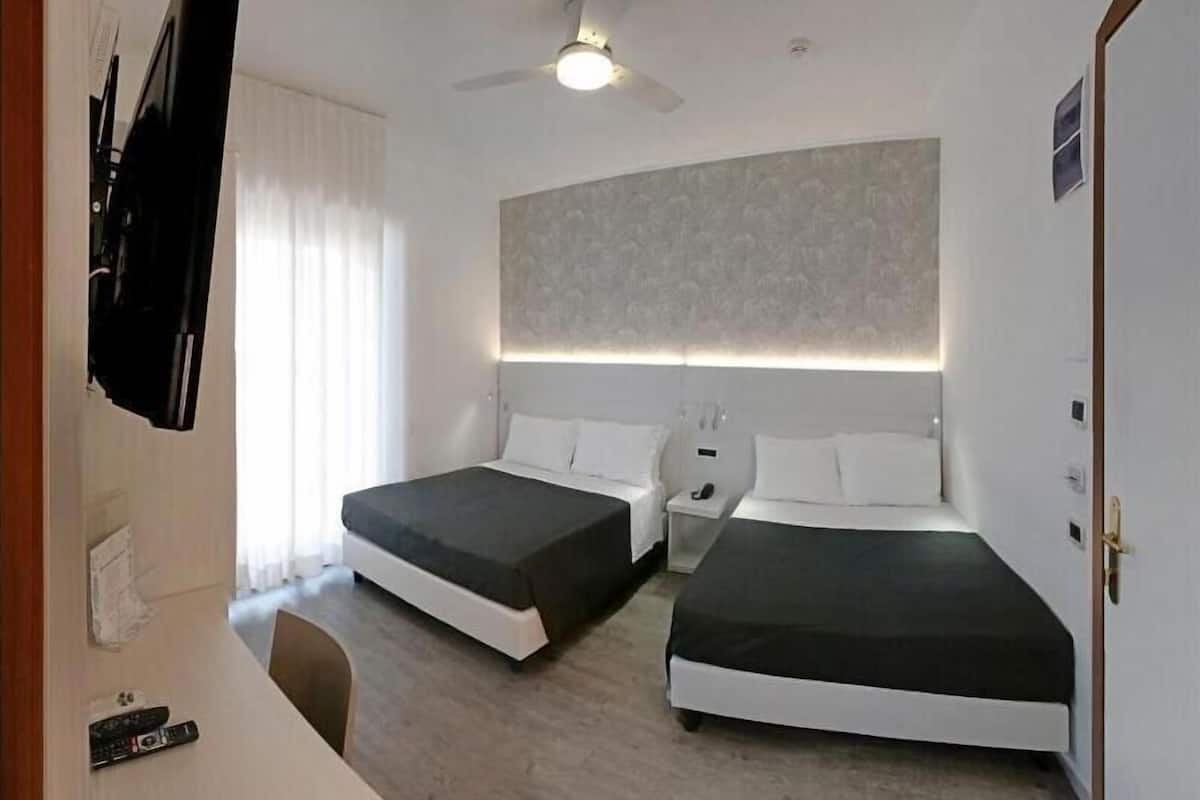 A hotel room with two single beds, a desk with a chair, a ceiling fan, and a large window with curtains.