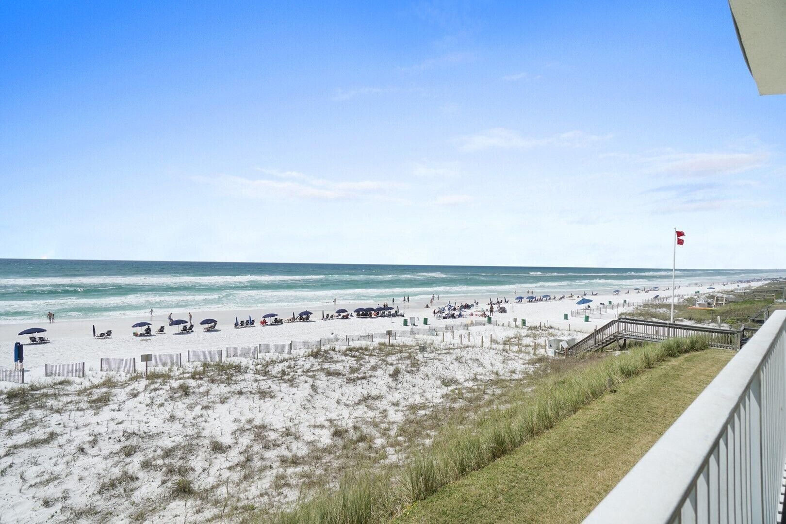 A beach with a wooden walkway leading to the shore, a flag, and people enjoying the beach.
