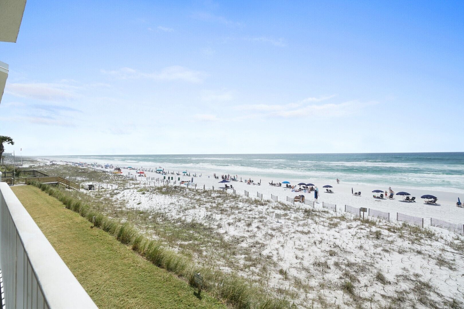 A beach with a wooden fence, beach chairs, umbrellas, and a grassy area.