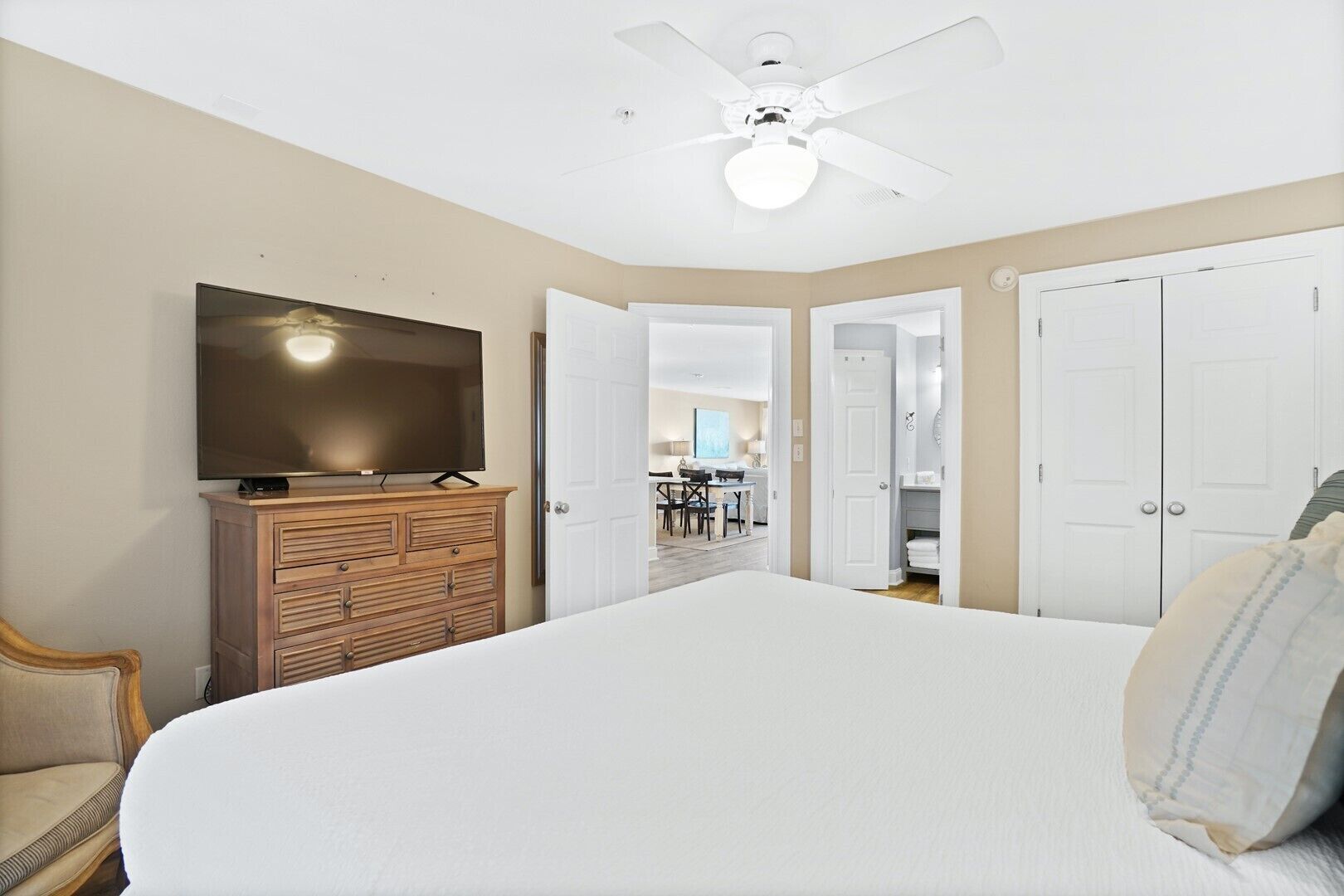 A bedroom with a large bed, a TV on a wooden dresser, a ceiling fan, and a view into the dining area.