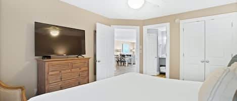 A bedroom with a large bed, a TV on a wooden dresser, a ceiling fan, and a view into the dining area.