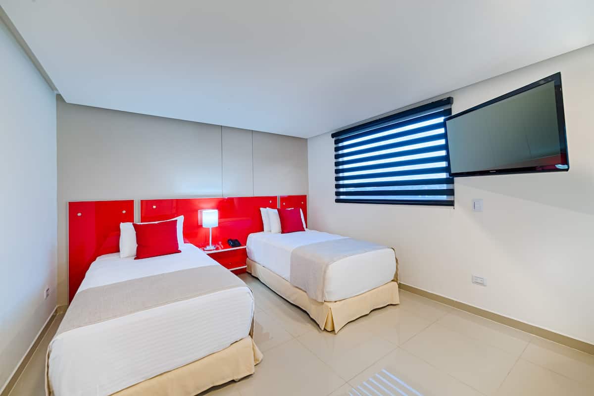 A hotel room with two beds, a red headboard, a flat-screen TV, and a window with blinds.
