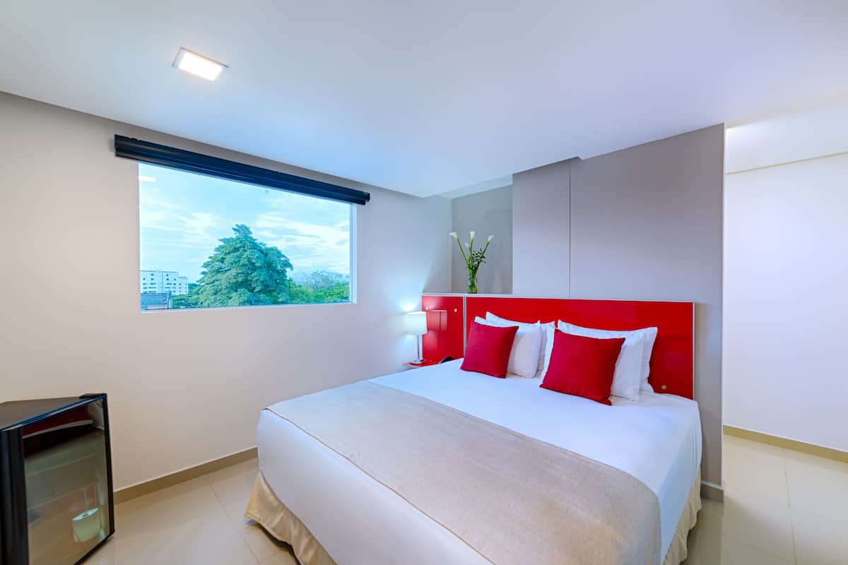 A modern hotel room with a large bed, a nightstand, a small table, and a window with a view of buildings and trees.