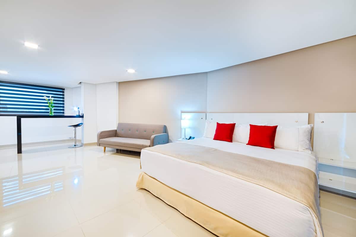 A modern hotel room with a large bed, a sofa, a desk, and a window with blinds.