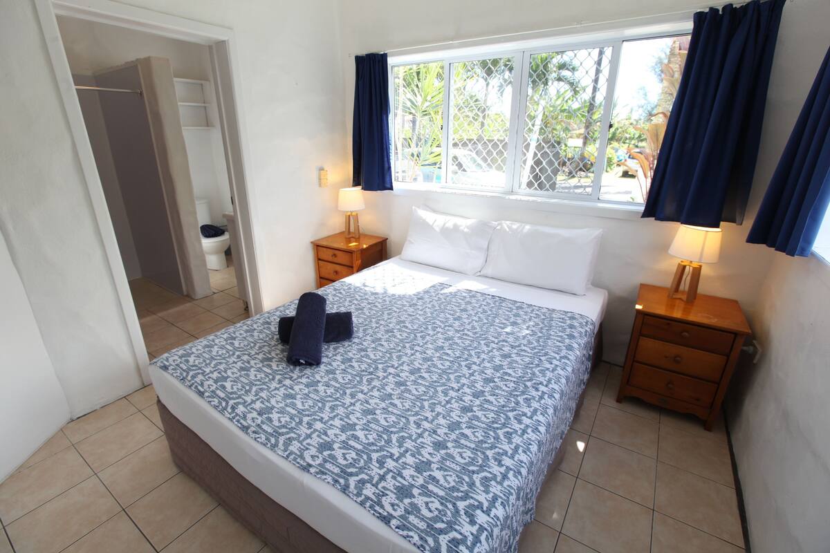 A bedroom with a bed, two bedside tables, a window with curtains, and a view of the outside.