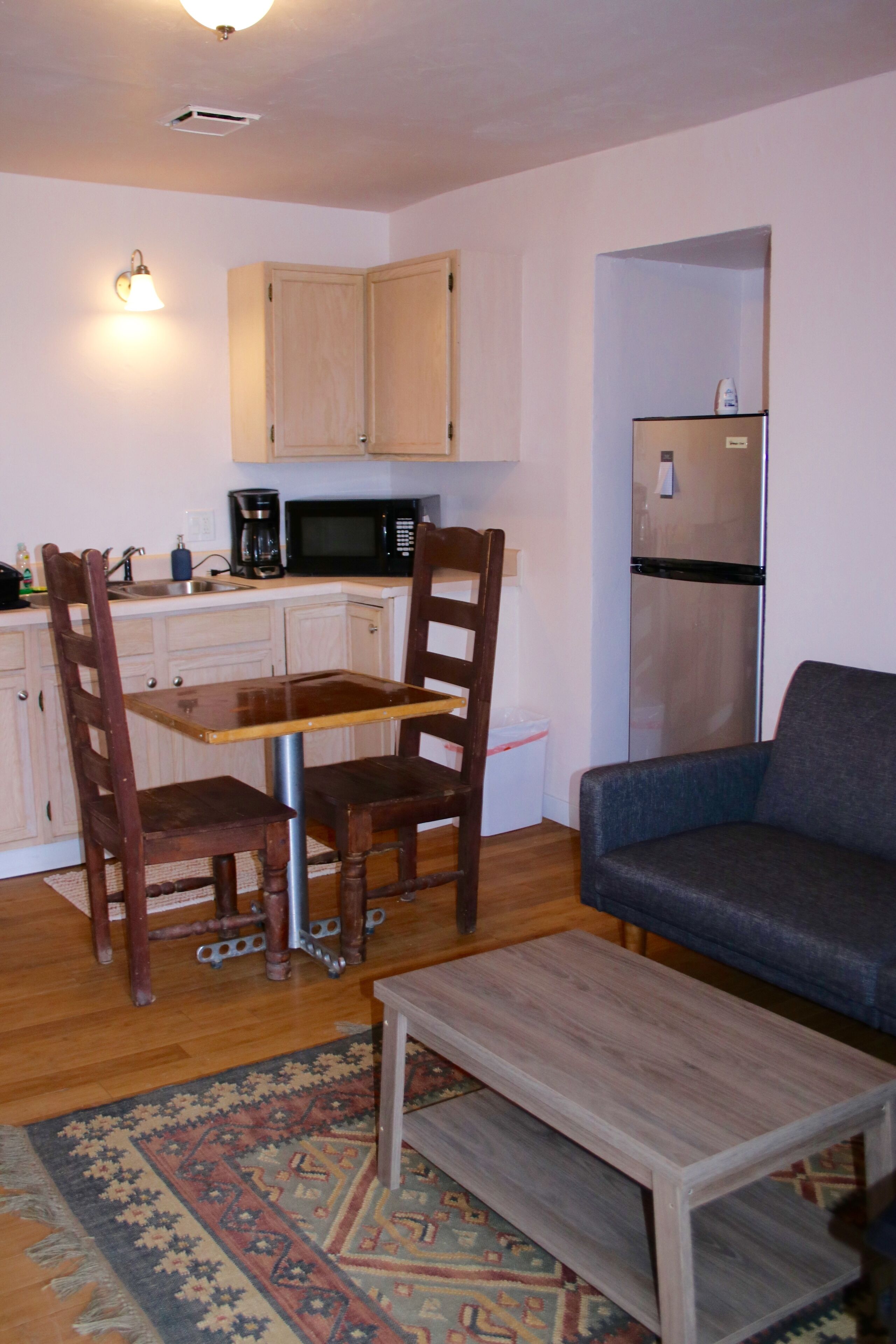 A compact living space with a dining table, two wooden chairs, a sofa, a small wooden coffee table, a kitchen area with a microwave, a refrigerator, and a patterned rug.