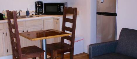 A compact living space with a dining table, two wooden chairs, a sofa, a small wooden coffee table, a kitchen area with a microwave, a refrigerator, and a patterned rug.