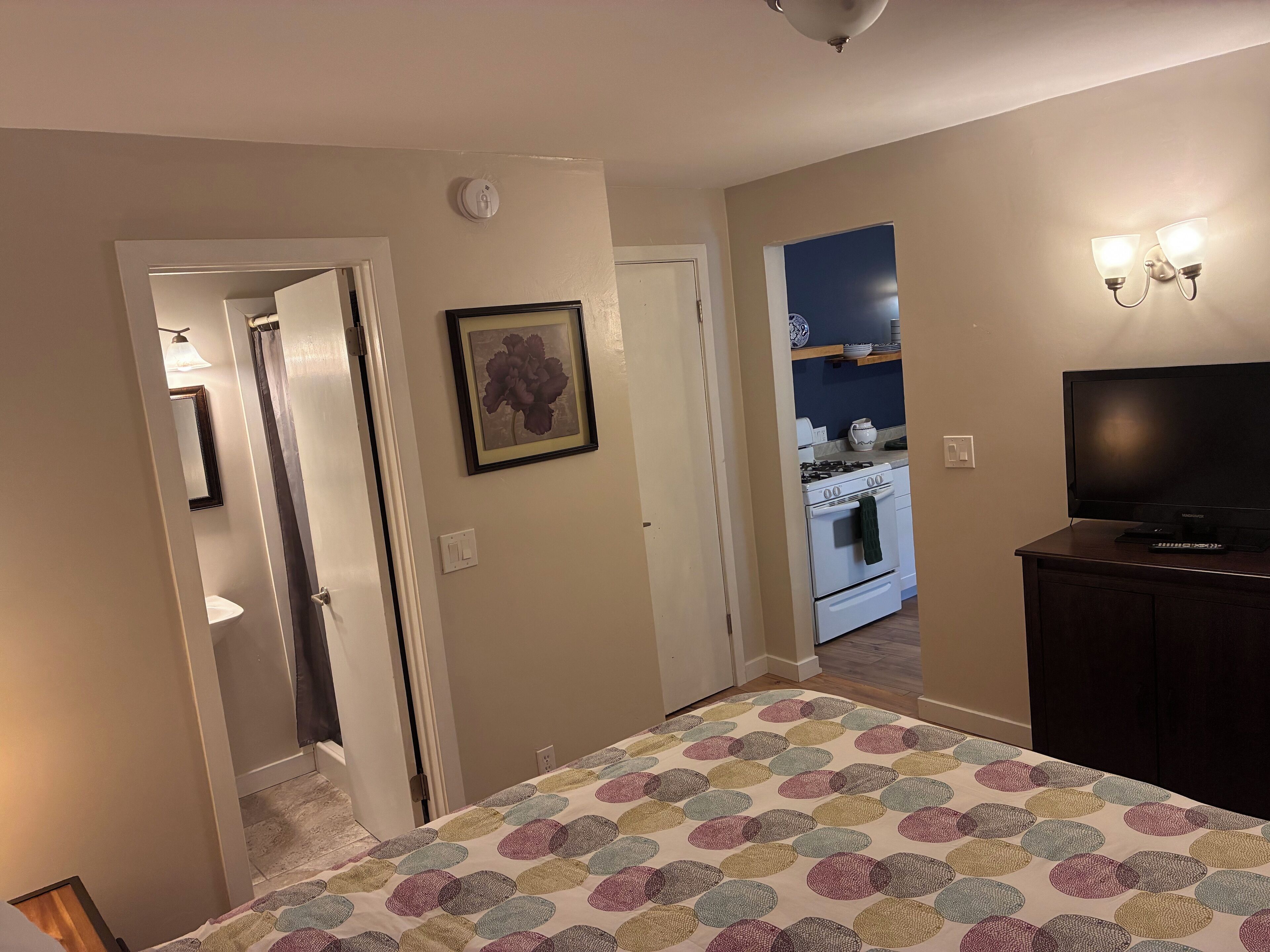 A bedroom with a bed, a TV, and a kitchen in the background.