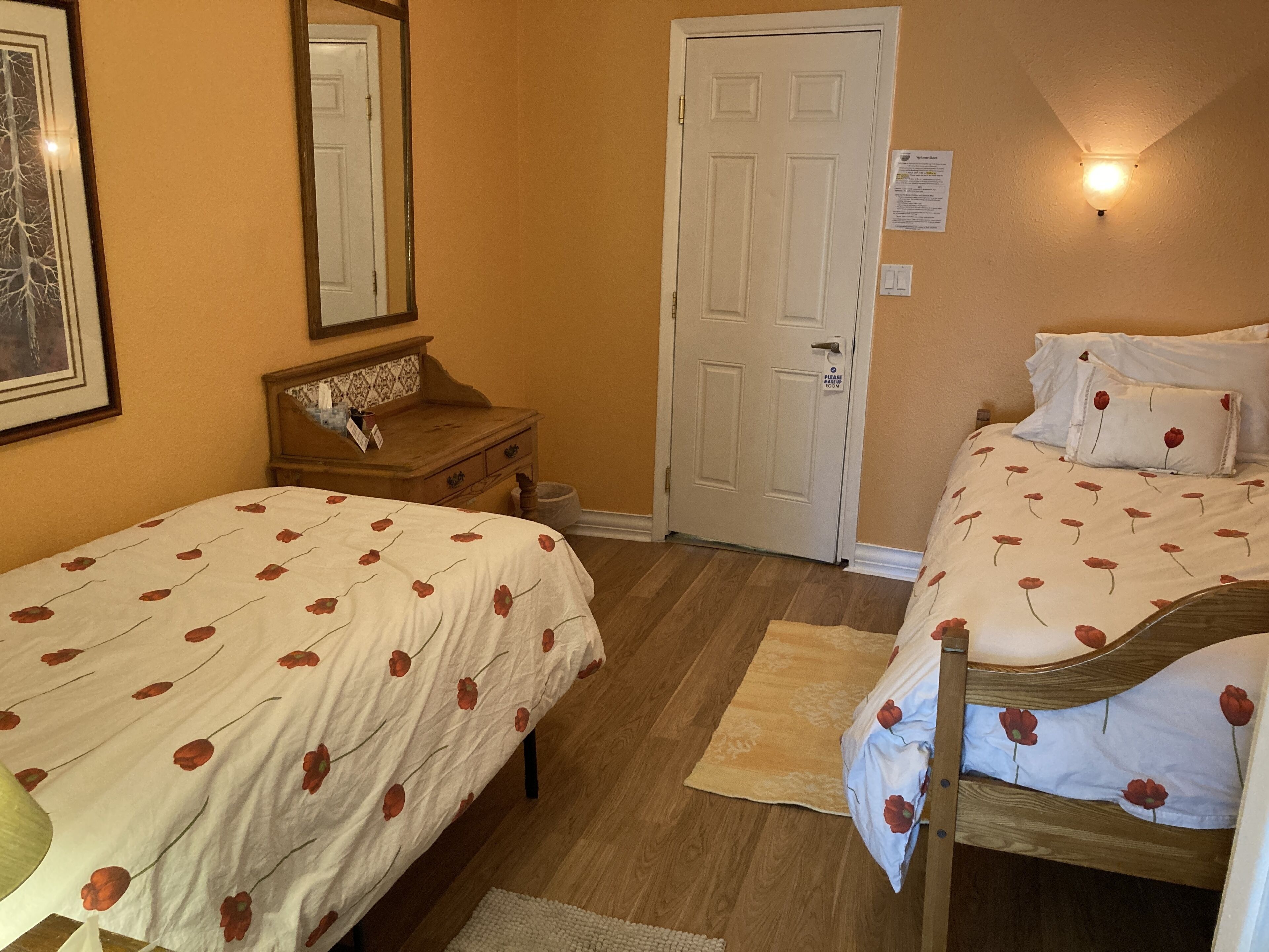 Een slaapkamer met twee eenpersoonsbedden, een houten dressoir, een spiegel, een deur en een wandlamp.