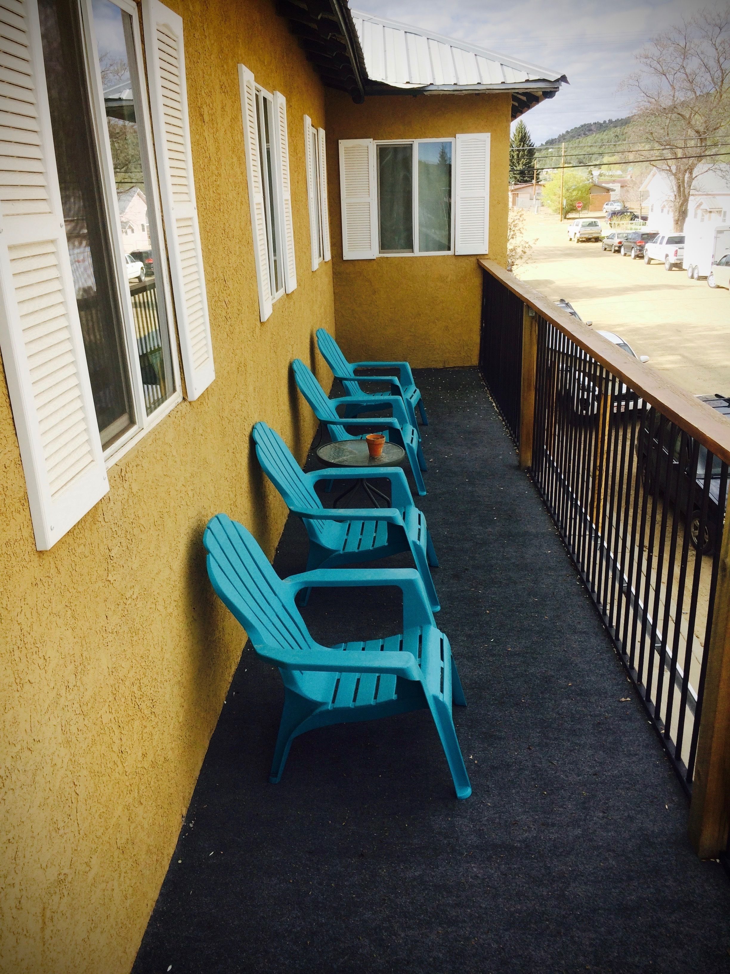 A balcony with blue plastic chairs and a small table, a yellow wall, and a view of parked cars and buildings in the background.