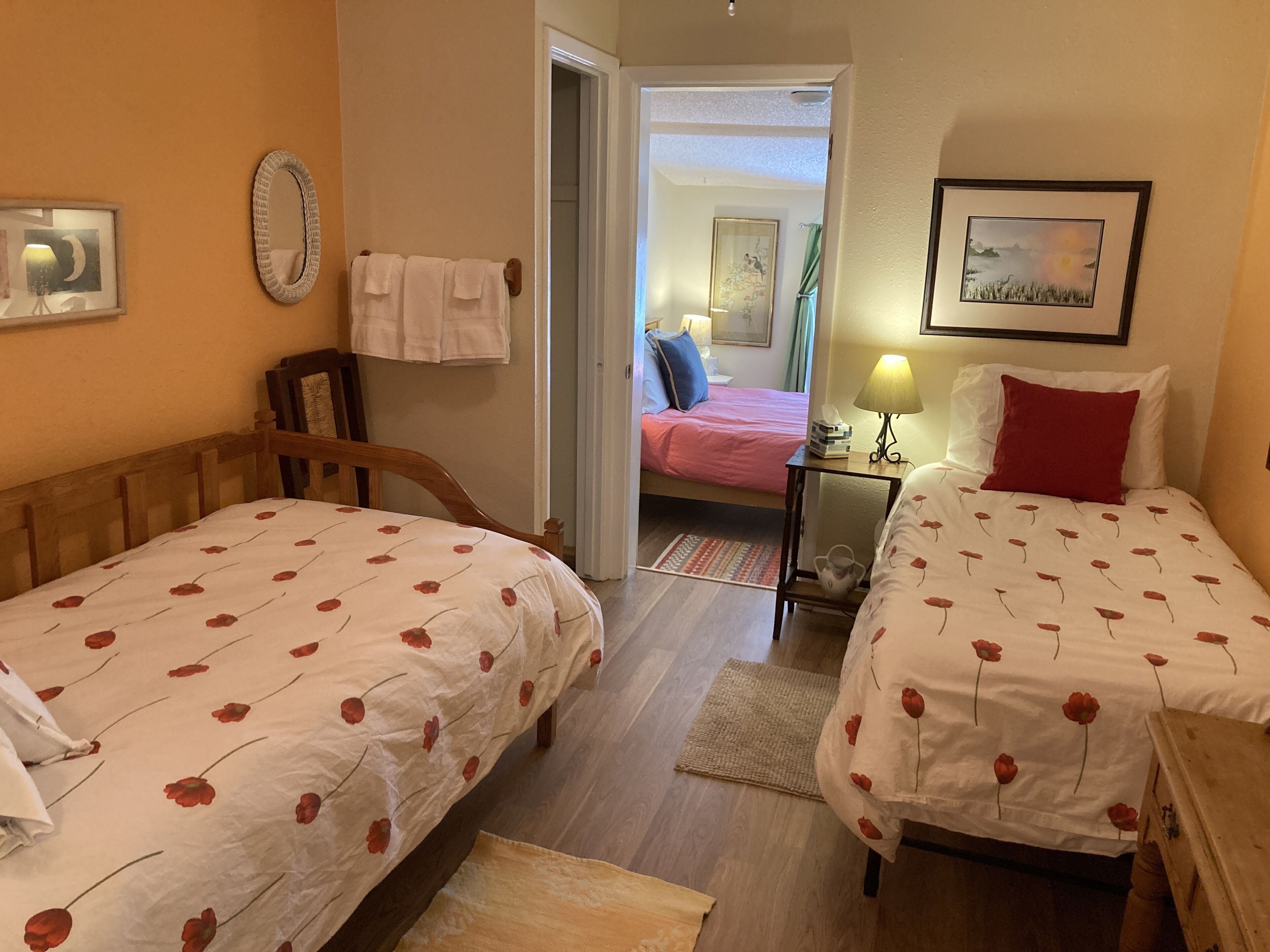 A hotel room with two single beds, a wooden headboard, a small table with a lamp, and a painting on the wall.
